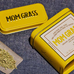Crushed CBG On A Rollin Paper With A Yellow Mom Grass CBG Hemp Flower Tin On A Grey Cloth