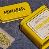 Crushed CBG On A Rollin Paper With A Yellow Mom Grass CBG Hemp Flower Tin On A Grey Cloth