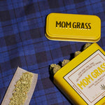 Crushed CBG On A Rollin Paper With A Yellow Mom Grass CBG Hemp Flower Tin On A Blue Cloth