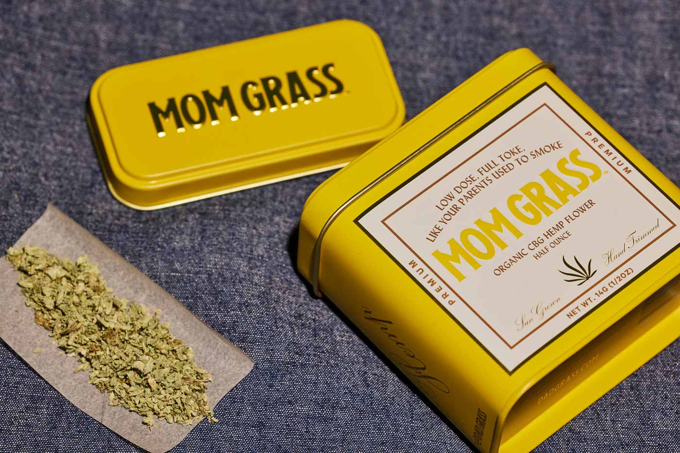 Crushed CBG On A Rollin Paper With A Yellow Mom Grass CBG Hemp Flower Tin On A Grey Cloth
